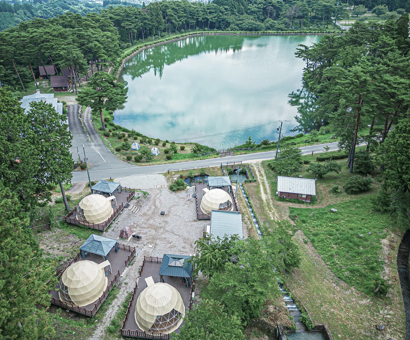湖が見える森の中に、グランピング用の白いドームテントが複数並んでいる風景。