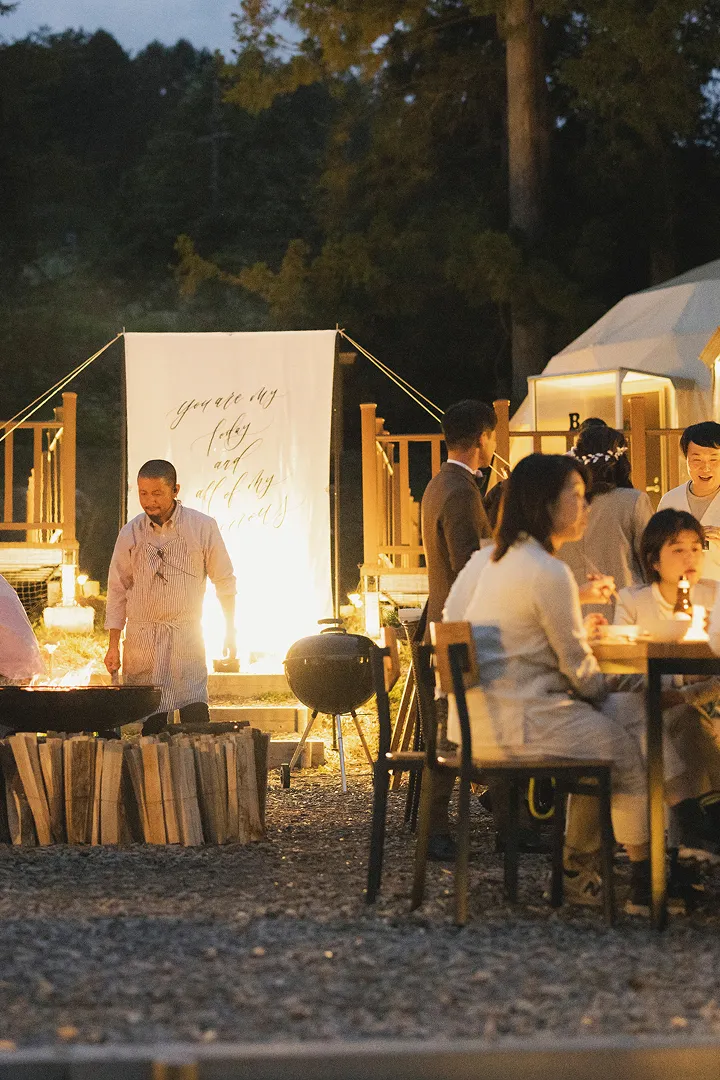 夜の屋外イベントで、ライトアップされたテントの前に人々が集まっている様子。