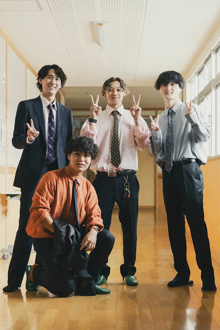 屋外でスーツやジャケットを着た4人の男性が、楽しそうにポーズをとっている集合写真。